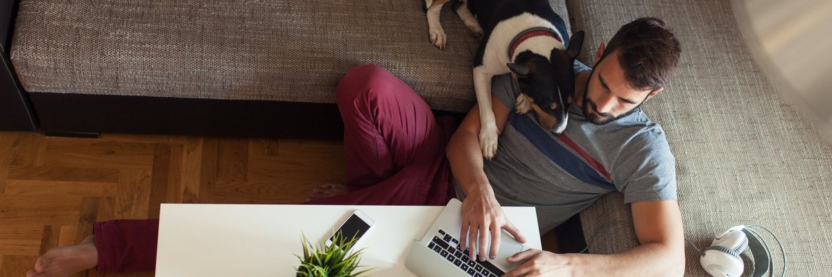 A man sitting on the floor in front of his couch with his dog looking over his shoulder at his open laptop.