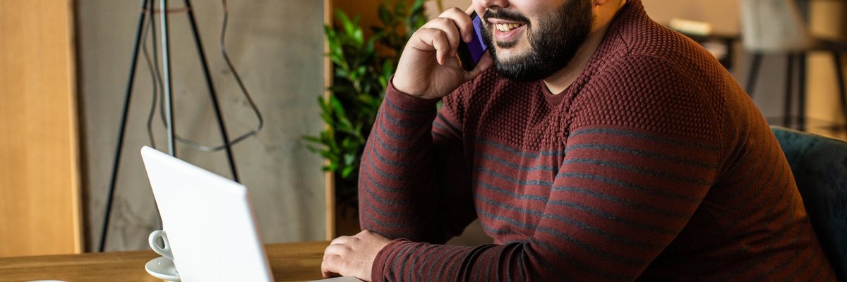A man talking on the phone while sitting in front of his laptop at a coffeeshop.