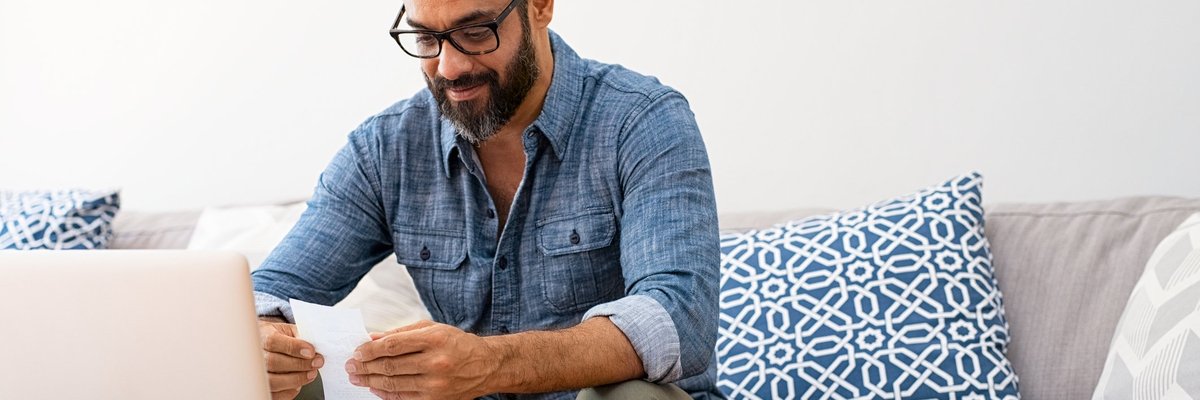 A man looking through paper bills while sitting on a couch in front of a laptop.