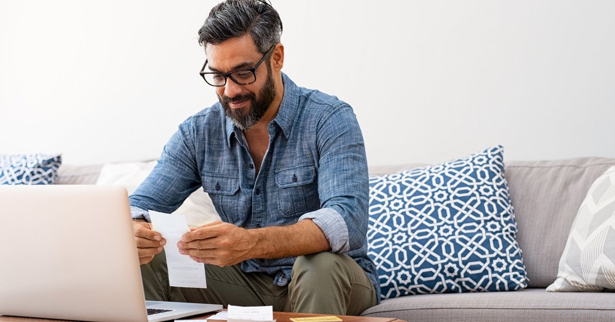 A man looking through paper bills while sitting on a couch in front of a laptop.