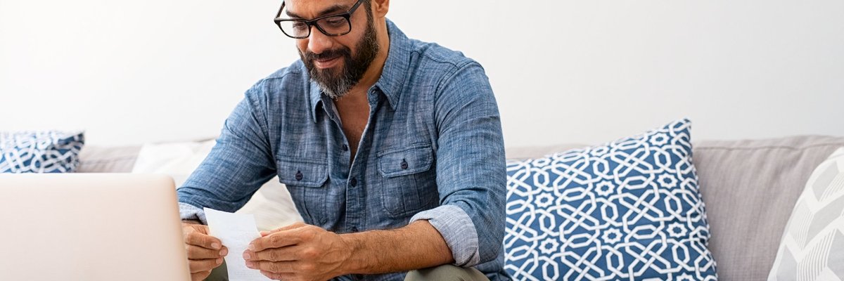 A man looking through paper bills while sitting on a couch in front of a laptop.