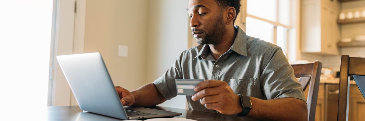 A man sits at a table, holding a credit card and paying his bills online.