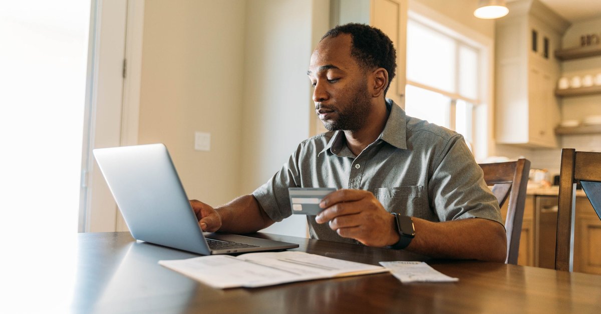 A man sits at a table, holding a credit card and paying his bills online.