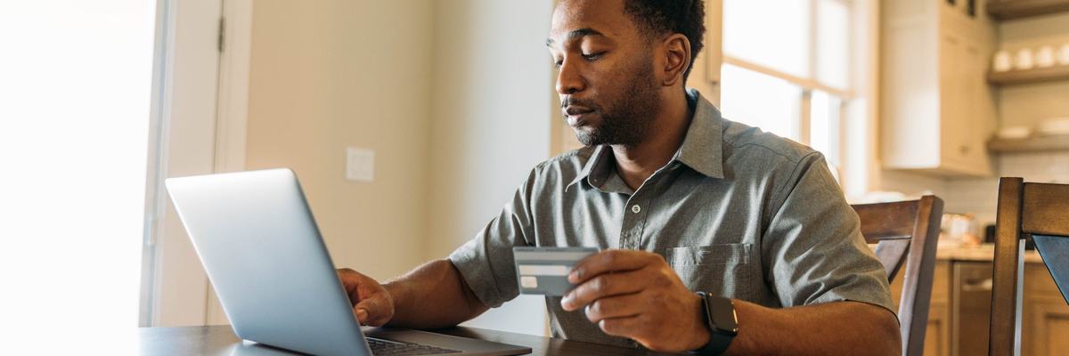 A man sits at a table, holding a credit card and paying his bills online.