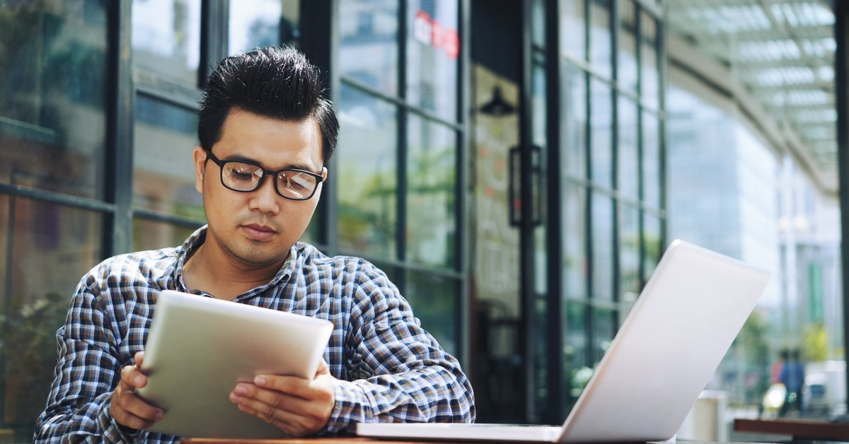 A man reading on a tablet with a laptop open on the table.