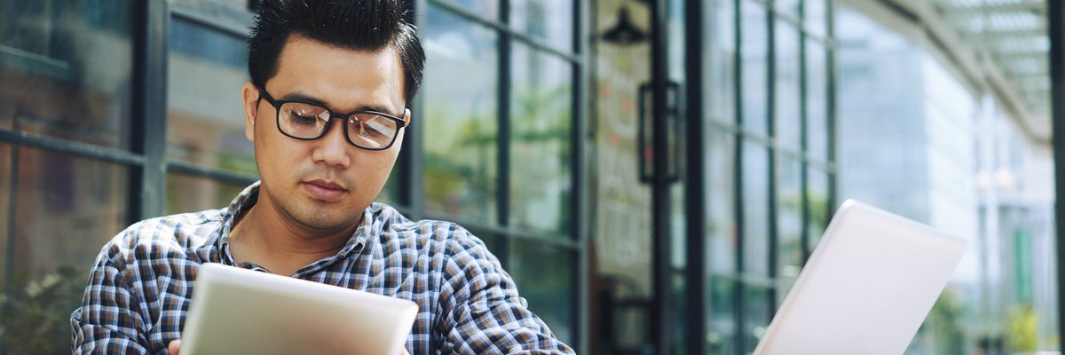 A man reading on a tablet with a laptop open on the table.