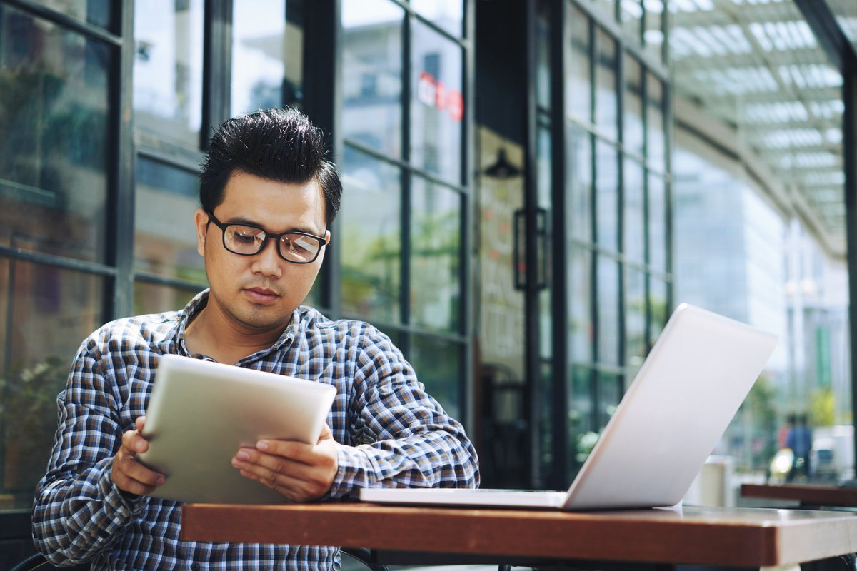 A man reading on a tablet with a laptop open on the table.