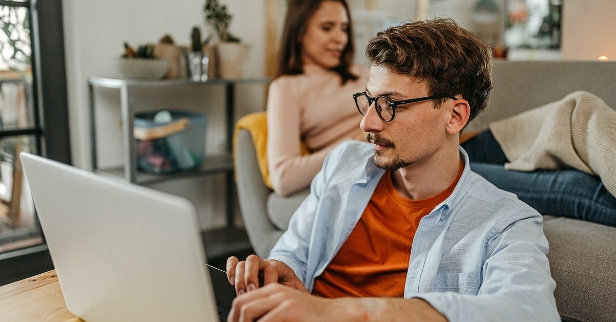 Man uses laptop while another person sits behind him on the couch.