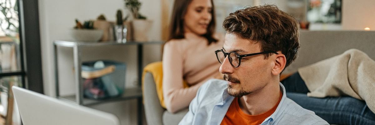 Man uses laptop while another person sits behind him on the couch.