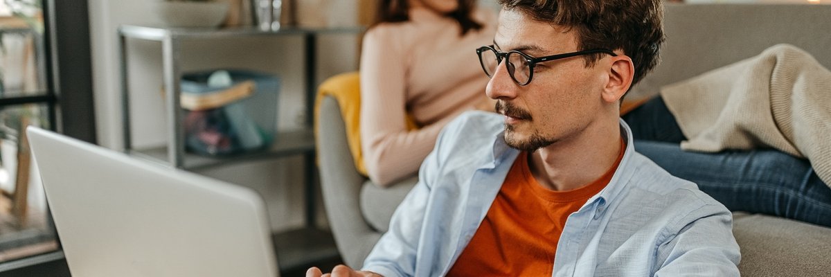 Man uses laptop while another person sits behind him on the couch.