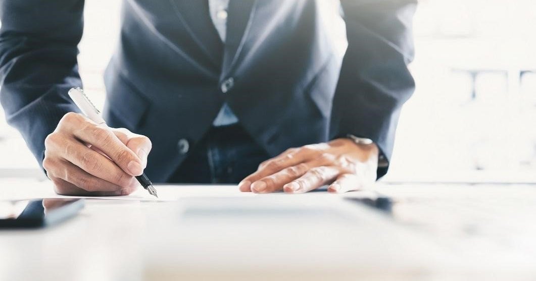 Man in suit signing paperwork on desk.