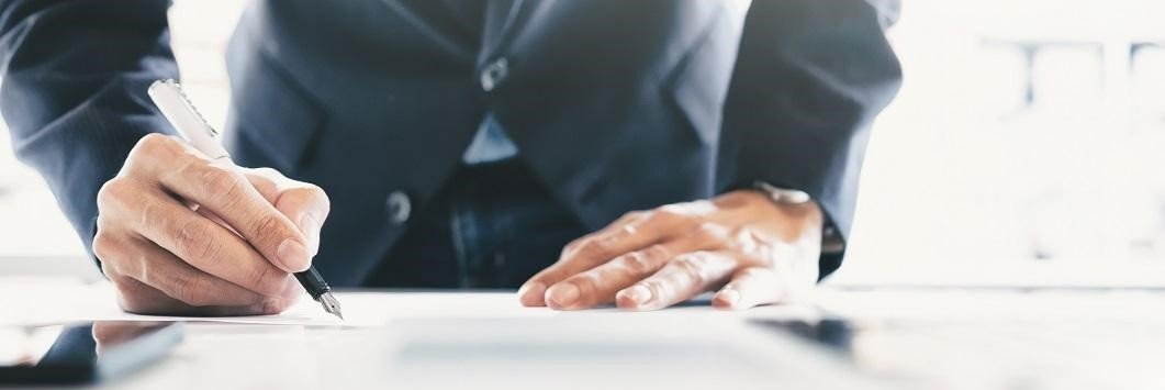 Man in suit signing paperwork on desk.