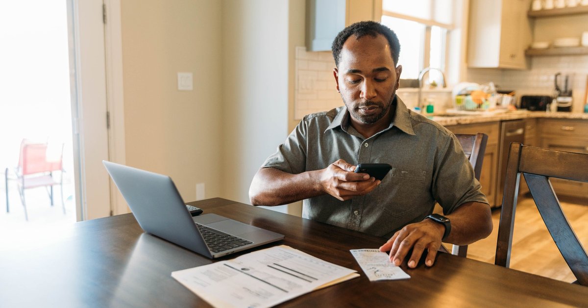 Man sits at counter with phone on laptop utilizing online banking.
