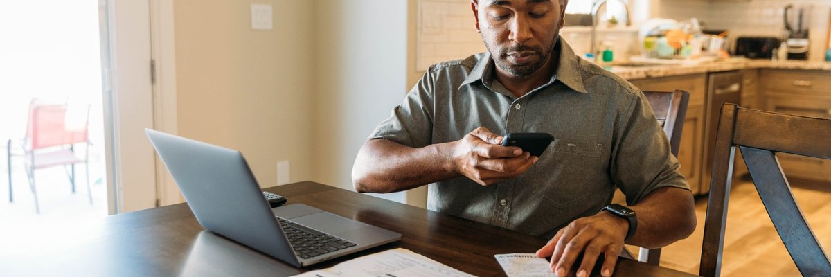 Man sits at counter with phone on laptop utilizing online banking.