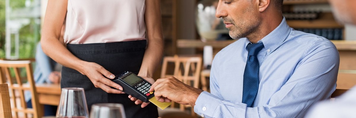 A man sitting in a restaurant swiping his credit card in the card reader being held by a waitress.