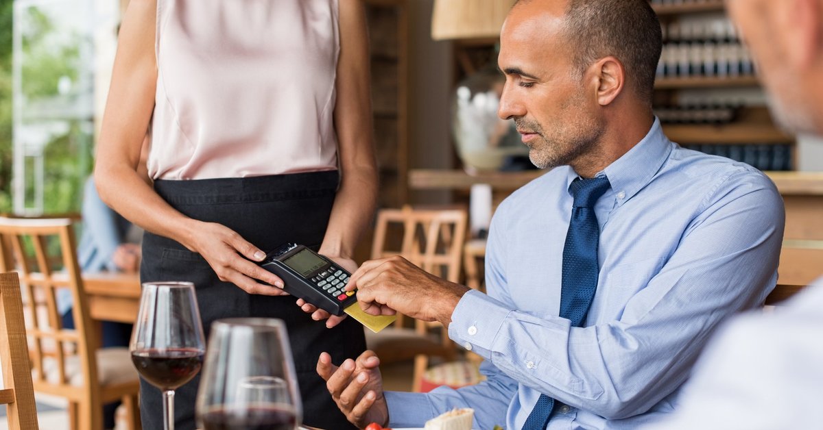 A man sitting in a restaurant swiping his credit card in the card reader being held by a waitress.