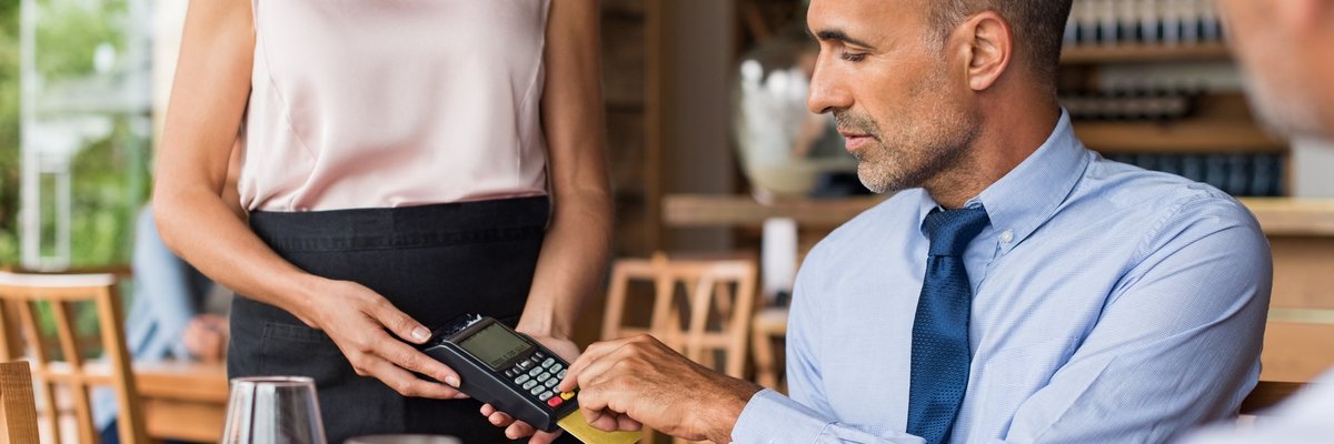 A businessman sitting at a restaurant table and swiping his credit card in the card reader being held by the waitress.