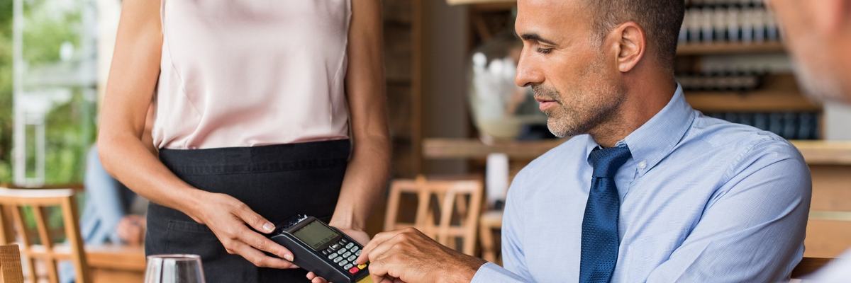 A man sitting in a restaurant swiping his credit card in the card reader being held by a waitress.