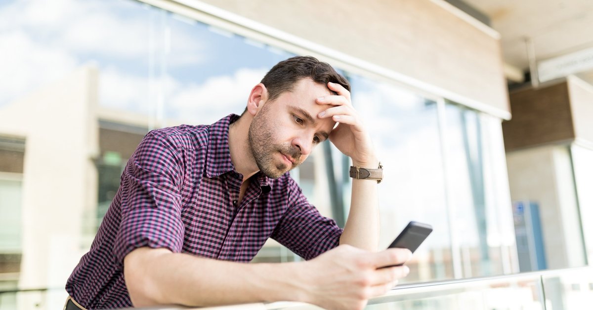 A man looking upset and leaning against a balcony railing while looking at his phone with his head resting in one hand.