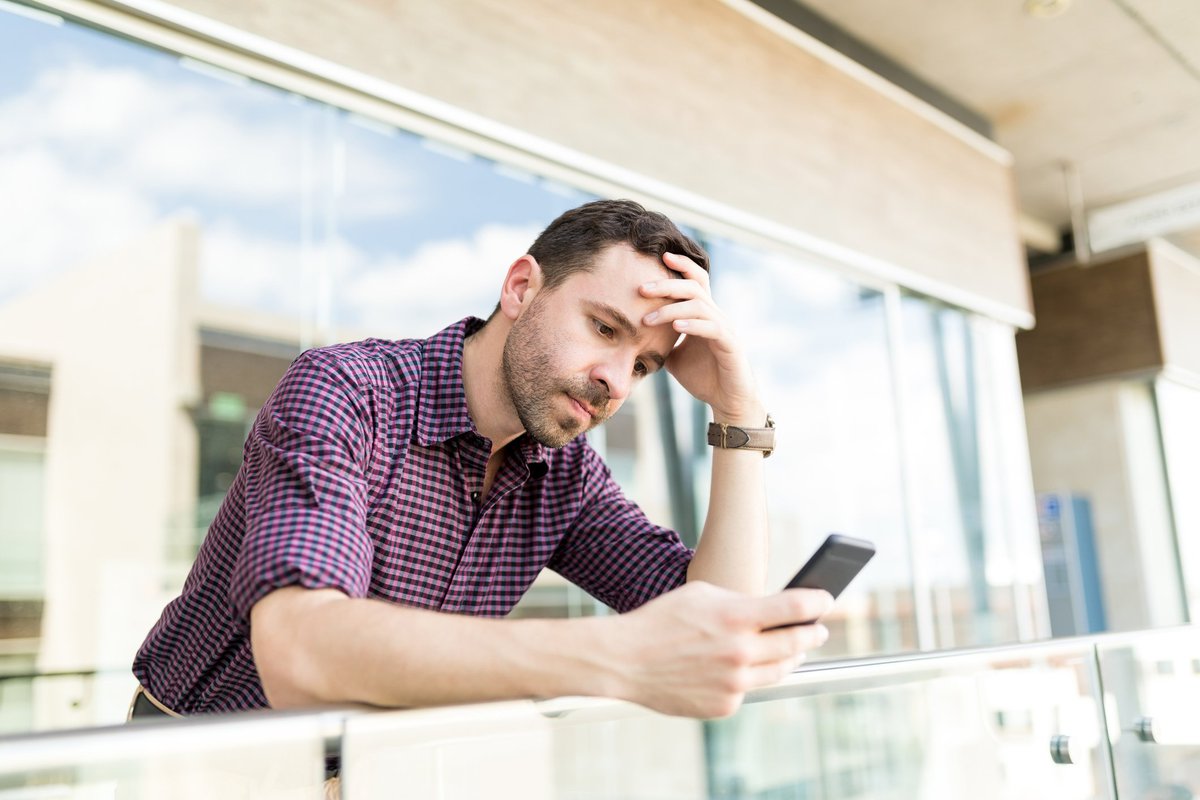 A man looks upset and leans up against a balcony railing while staring at his phone with his head resting in one hand.