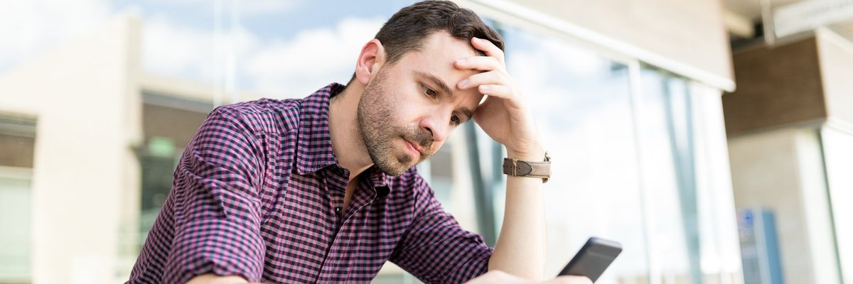 A man looking upset and leaning against a balcony railing while looking at his phone with his head resting in one hand.