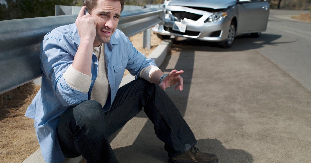 Man using his cellphone on the side of the road with a wrecked vehicle behind him.