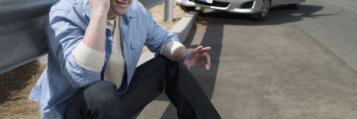 Man using his cellphone on the side of the road with a wrecked vehicle behind him.