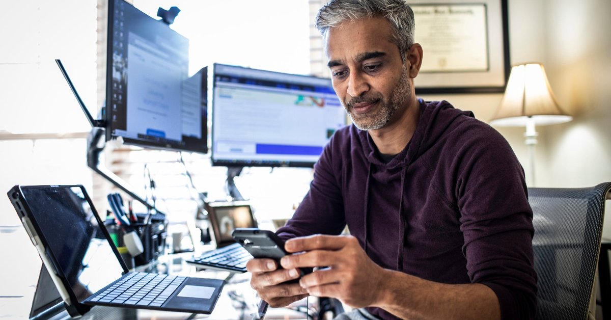 Man using is cell phone while sitting at desk with laptop and dual monitors.