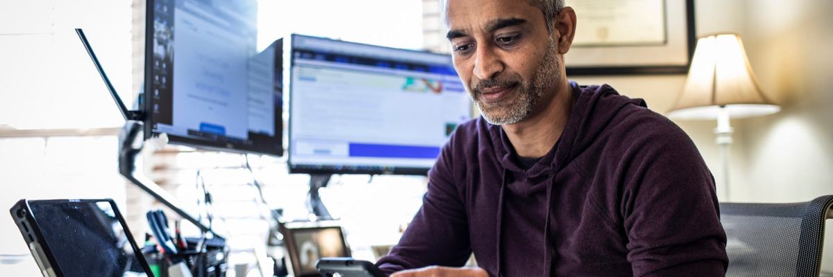 Man using is cell phone while sitting at desk with laptop and dual monitors.