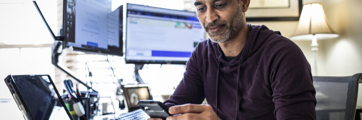 Man using is cell phone while sitting at desk with laptop and dual monitors.