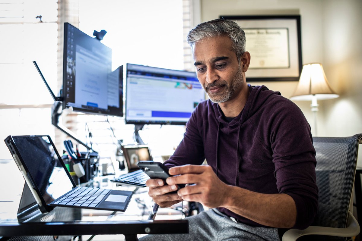 Man using is cell phone while sitting at desk with laptop and dual monitors.