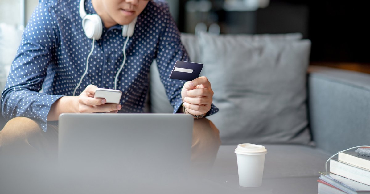 A man sitting on his couch in front of a laptop while holding his phone and a credit card.