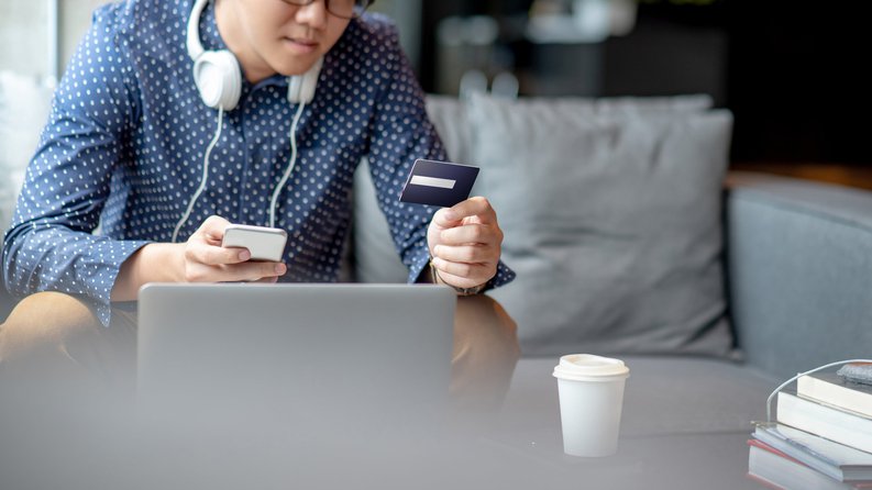 A man sitting on his couch in front of a laptop while holding his phone and a credit card.