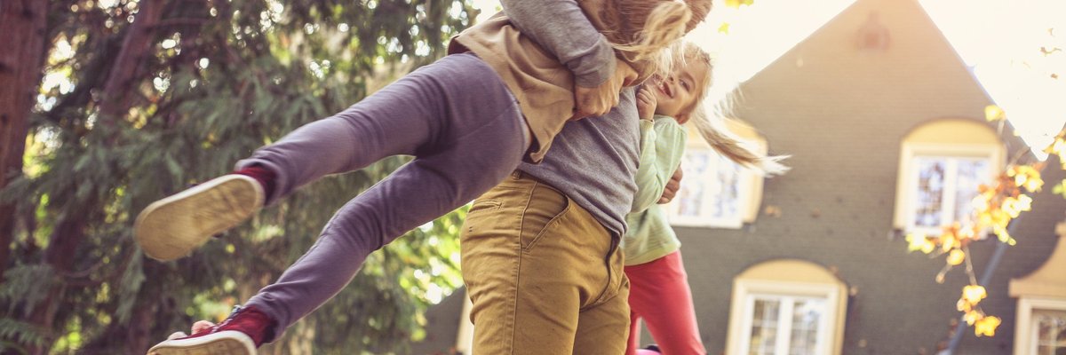 A laughing man swinging his daughters in the air in their backyard.