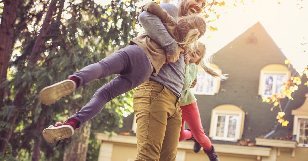 A laughing man swinging his daughters in the air in their backyard.