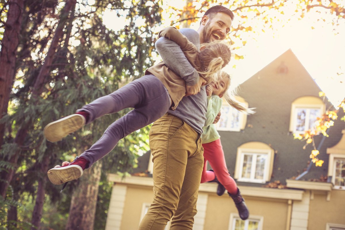 A laughing man swinging his daughters in the air in their backyard.