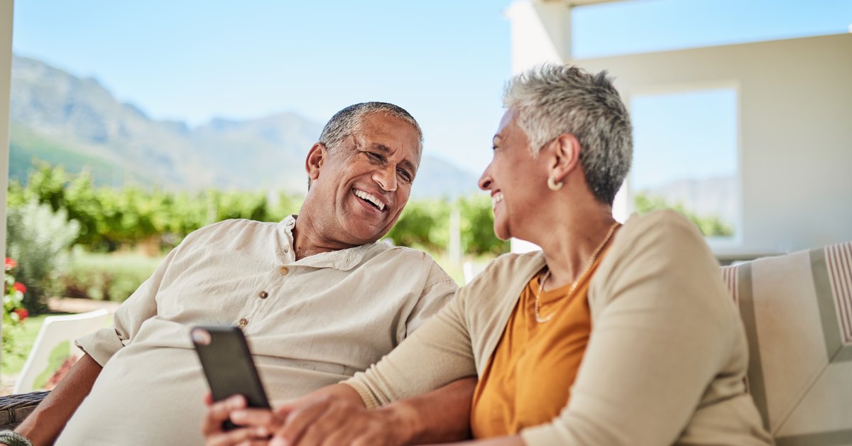 Mature couple smiling with cellphone in hand while sitting outdoors among background of mountains