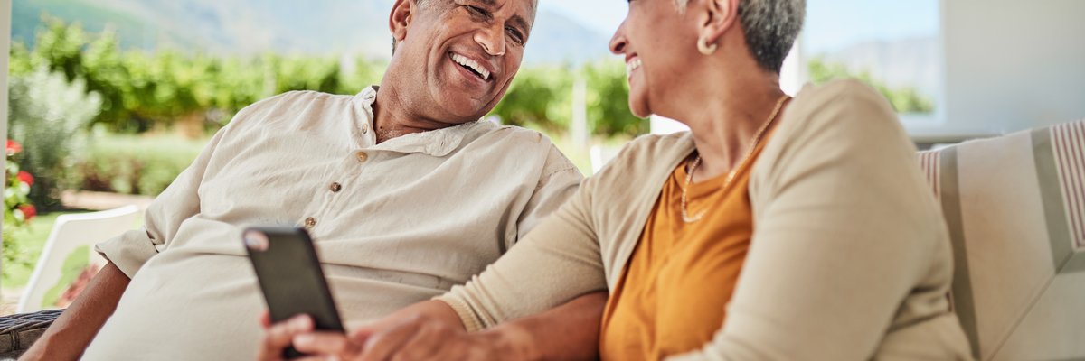 Mature couple smiling with cellphone in hand while sitting outdoors among background of mountains