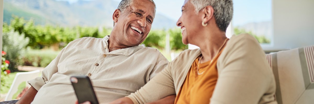 Mature couple smiling with cellphone in hand while sitting outdoors among background of mountains