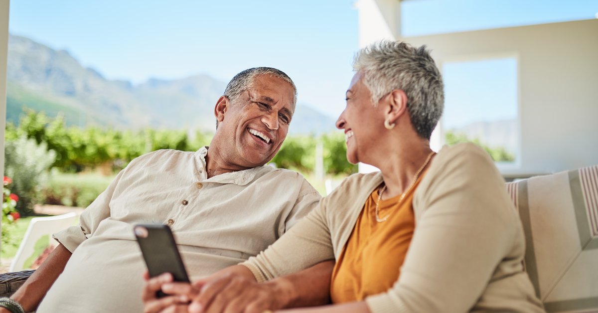 Mature couple smiling with cellphone in hand while sitting outdoors among background of mountains