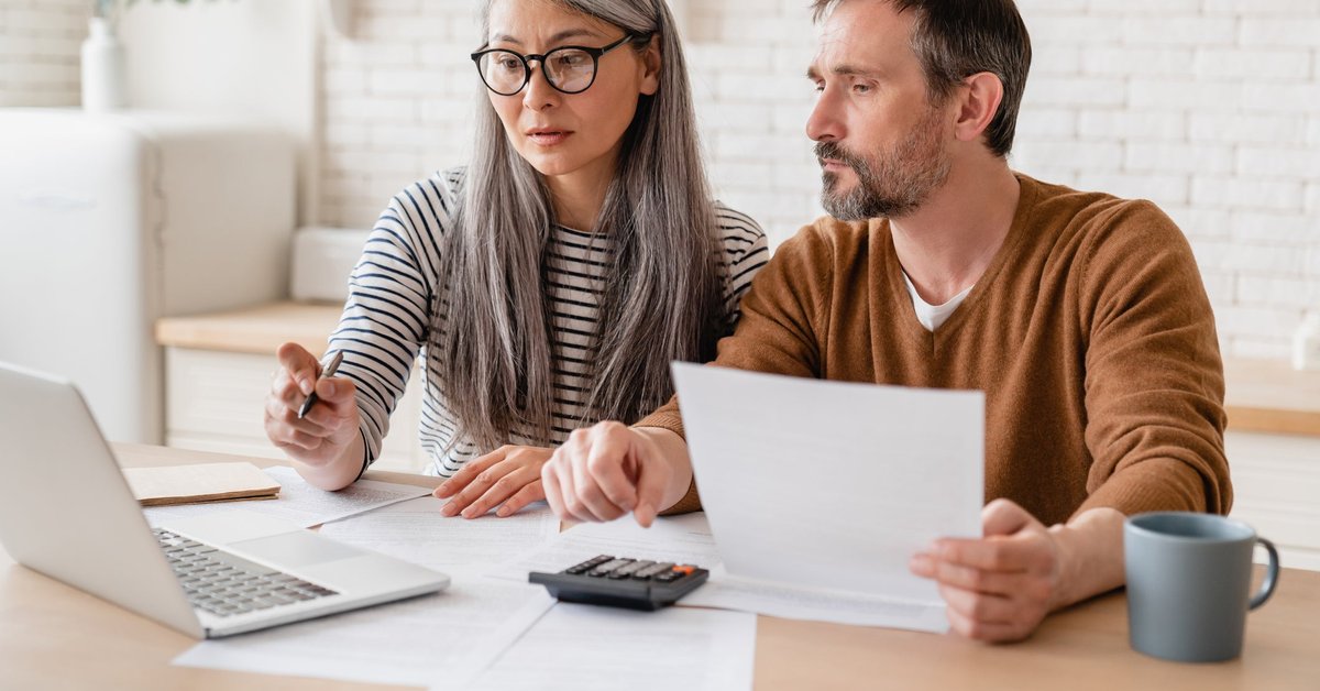 A mature couple sitting at a kitchen table with papers, a laptop, and a calculator.