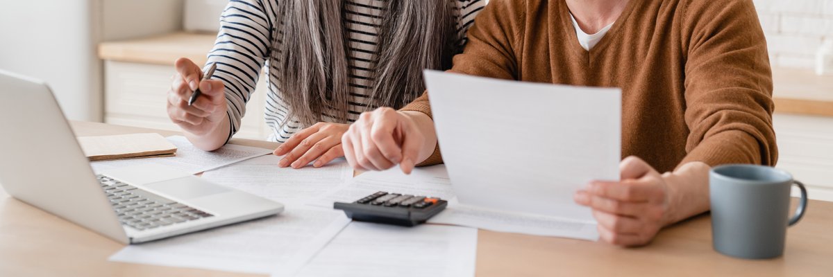 A mature couple sitting at a kitchen table with papers, a laptop, and a calculator.