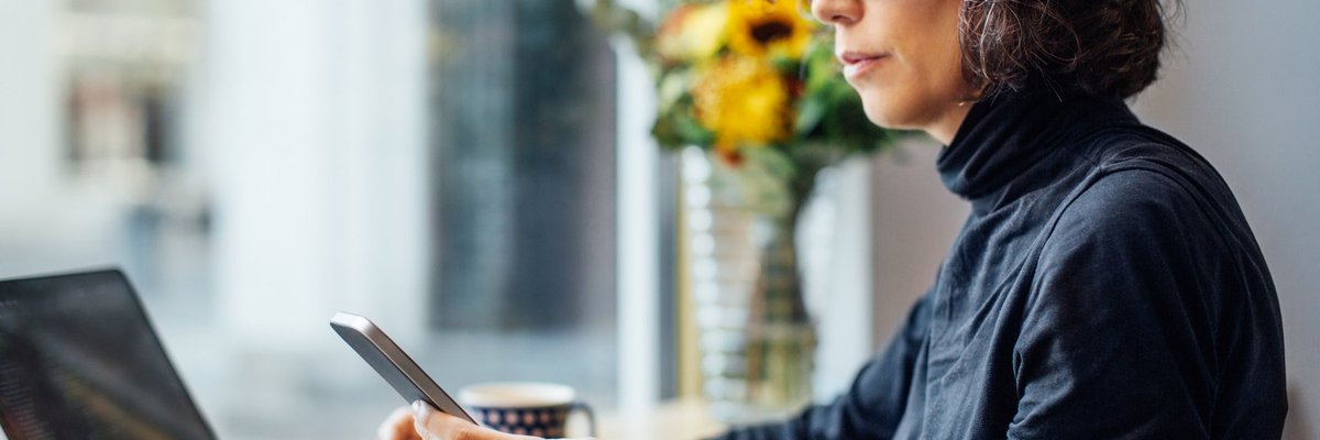 Mature women looking at her cell phone with a laptop in front of her.