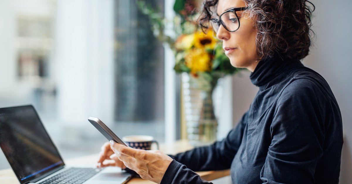 Mature women looking at her cell phone with a laptop in front of her.
