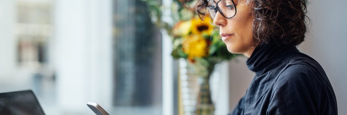Mature women looking at her cell phone with a laptop in front of her.