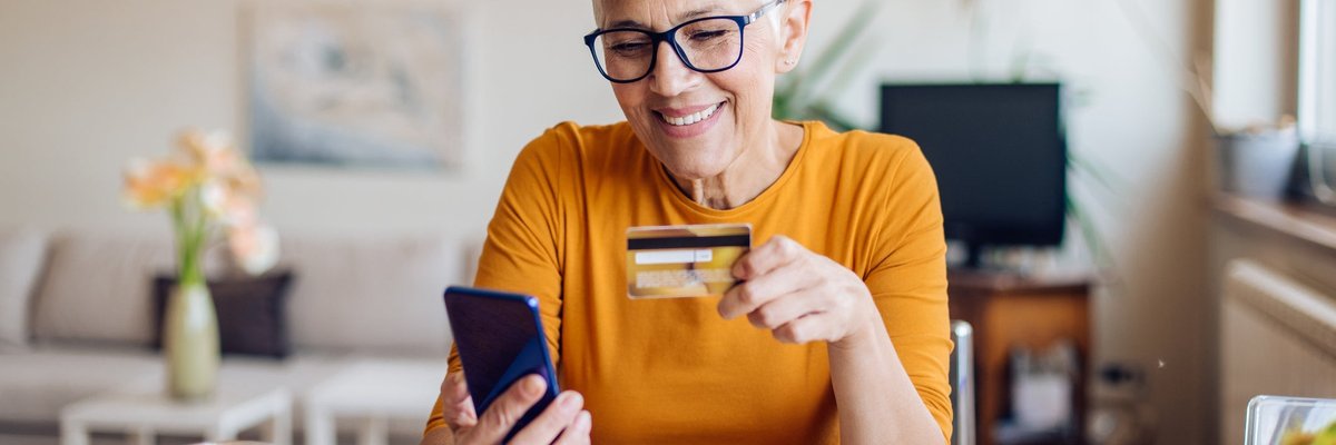 A mature woman smiles while sitting at her table. She is holding a cellphone and a credit card.