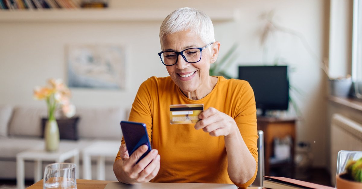 A mature woman smiles while sitting at her table. She is holding a cellphone and a credit card.