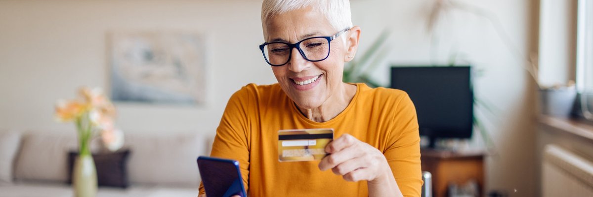 A mature woman smiles while sitting at her table. She is holding a cellphone and a credit card.