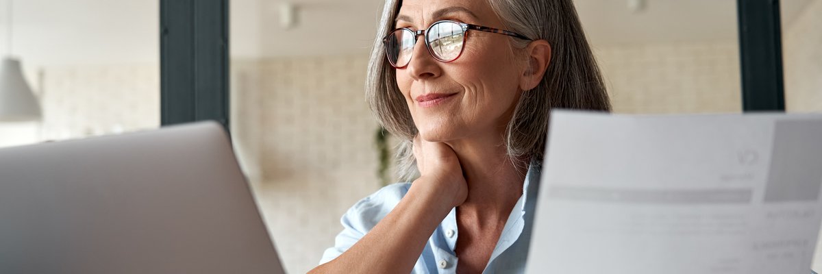 Mature woman working from laptop with documents in hand.