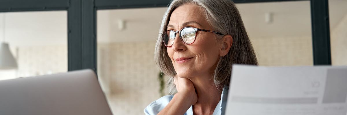 Mature woman working from laptop with documents in hand.